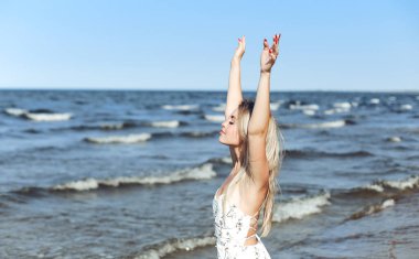 Okyanus sahilinde beyaz bir yaz elbisesi içinde elini kaldıran mutlu sarışın güzel kadın..