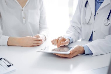 Doktor ve hasta klinikteki masada yan yana oturuyorlar. Kadın doktorların elleri tablet bilgisayarın dokunmatik dokunmatik pedini işaret ediyor, kapat. Tıp konsepti.