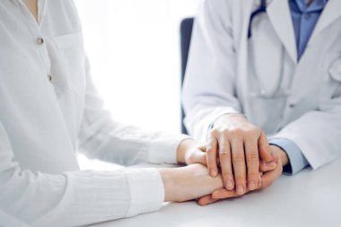 Doctor and patient sitting near each other at the table in clinic office. The focus is on female physicians hands reassuring woman, only hands, close up. Medicine concept. clipart