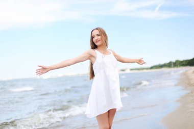 Okyanus sahilinde mutlu, mutlu ve mutlu bir kadın elleri açık duruyor. Beyaz yaz elbisesi içinde çok kültürlü bir kadın modelin portresi seyahat tatilinde doğanın tadını çıkarıyor.