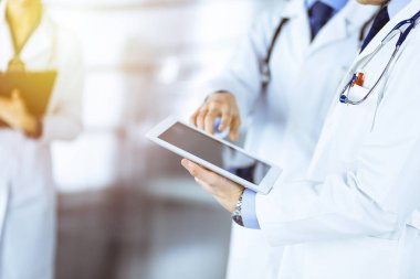 Bilinmeyen bir grup doktor, güneşli bir hastane ofisinde dururken bazı tıbbi isim kayıtlarını kontrol etmek için bir bilgisayar tableti kullanıyor. Doktorlar hastaları muayene etmeye ve yardım etmeye hazır. Tıbbi yardım, sigorta.