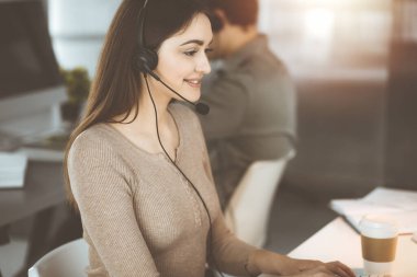 Kulaklık takmış genç bir kız, güneşli bir ofiste masada otururken bir firma müşterisiyle konuşuyor. İş yerindeki çağrı merkezi operatörleri.