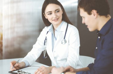 Dost canlısı genç bayan doktor, güneşli klinikte birlikte otururken hastasını dinliyor. Tıp ve sağlık hizmetleri kavramları.