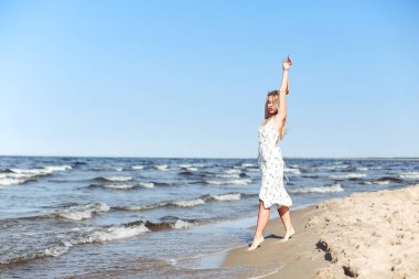 Okyanus sahilinde beyaz bir yaz elbisesi içinde kollarını açmış mutlu sarışın güzel bir kadın..
