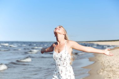 Okyanus sahilinde beyaz bir yaz elbisesi içinde elini kaldıran mutlu sarışın güzel kadın..