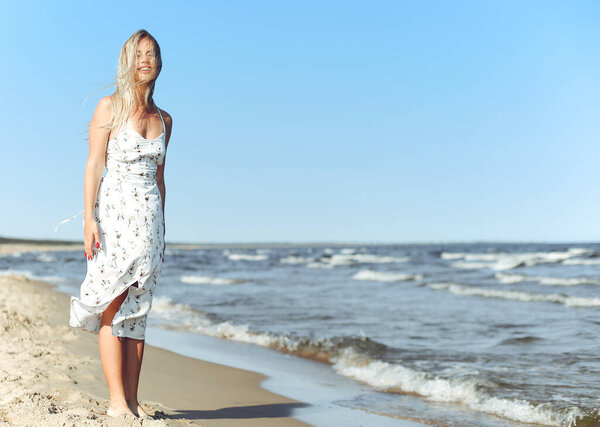 Счастливая блондинка в свободном счастье блаженство на берегу океана стоя прямо. Портрет женщины-модели в белом летнем платье, наслаждающейся природой во время отпуска на природе.