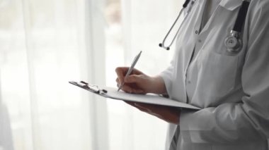 Doktor kadın pano tutarken ve klinikteki panorama penceresinin yanında dururken tıbbi kayıtları dolduruyor. Tıp konsepti.