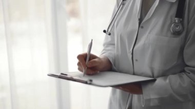Doktor kadın pano tutarken ve klinikteki panorama penceresinin yanında dururken tıbbi kayıtları dolduruyor. Tıp konsepti.