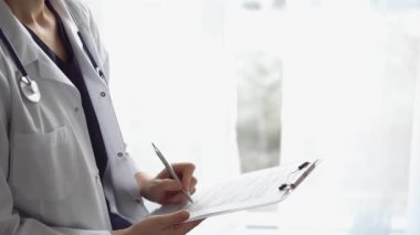 Doktor kadın, tıbbi notlar yazarken beyaz bir not panosu kullanıyor. Hastanenin panorama penceresinin yanında duran bilinmeyen bir doktor. İlaç..