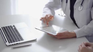 Doktor ve hasta klinikteki özel pencerenin yanındaki beyaz masada oturuyorlar. Beyaz önlük giyen kimliği belirsiz kadın doktor tıbbi kayıt formlarını doldurmak için tablet bilgisayar kullanıyor. Tıp konsepti.