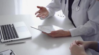 Doktor ve hasta klinikteki özel pencerenin yanındaki beyaz masada oturuyorlar. Beyaz önlük giyen kimliği belirsiz kadın doktor tıbbi kayıt formlarını doldurmak için tablet bilgisayar kullanıyor. Tıp konsepti.