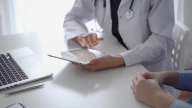 Doktor ve hasta klinikteki özel pencerenin yanındaki beyaz masada oturuyorlar. Beyaz önlük giyen kimliği belirsiz kadın doktor tıbbi kayıt formlarını doldurmak için tablet bilgisayar kullanıyor. Tıp konsepti.