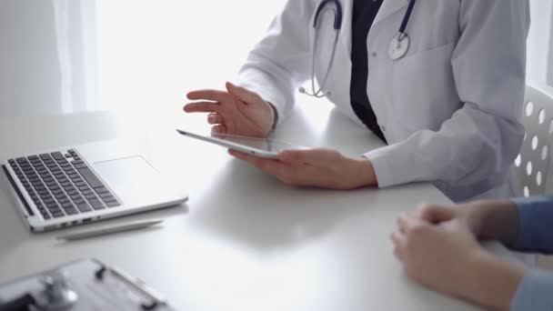 医生和病人坐在诊所亚麻窗附近的白桌前 穿着白衣的无名女医生用平板电脑填写病历表 医学概念 — 图库视频影像