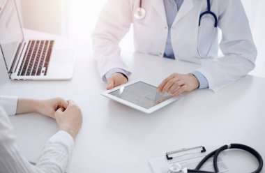 Doktor ve hasta klinikteki masada karşılıklı oturuyorlar. Kadın doktorların elleri tablet bilgisayarın dokunmatik dokunmatik pedini işaret ediyor, kapat. Tıp konsepti.