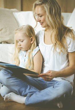 Mutlu bir aile. Sarışın genç anne ahşap zeminde otururken sevimli kızına kitap okuyor. Annelik kavramı.