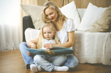 Mutlu bir aile. Sarışın genç anne ahşap zeminde otururken sevimli kızına kitap okuyor. Annelik kavramı.