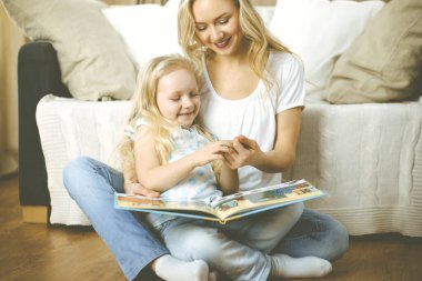 Mutlu bir aile. Sarışın genç anne ahşap zeminde otururken sevimli kızına kitap okuyor. Annelik kavramı.