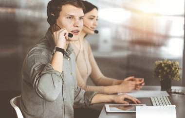 Call center operators at work. Two young people in headsets are talking to the clients, while sitting in sunny office. clipart