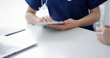 Doktor ve hasta klinikte oturup tablet bilgisayar kullanırken birbirleriyle konuşuyorlar, kapatın. Tıp konsepti.