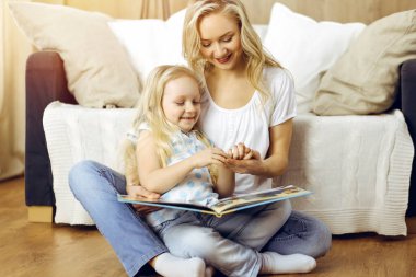 Mutlu bir aile. Sarışın genç anne güneşli odada ahşap zeminde otururken sevimli kızına kitap okuyor. Annelik kavramı.