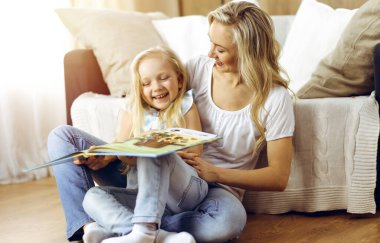 Mutlu bir aile. Sarışın genç anne güneşli odada ahşap zeminde otururken sevimli kızına kitap okuyor. Annelik kavramı.