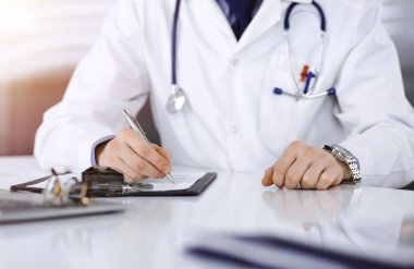 Bilinmeyen bir erkek doktor, karanlık bir klinikte tıbbi geçmişi olan bir panoyla oturuyor ve çalışıyor. Arka planda ışık parıltısı var..