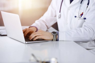 Bilinmeyen bir erkek doktor karanlık bir klinikte dizüstü bilgisayarla oturuyor ve çalışıyor. Arka planda ışık parıltısı var..