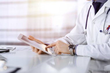 Bilinmeyen erkek doktor, tablet bilgisayarla oturuyor ve çalışıyor. Karanlık bir klinikte, arka planda ışık parıltısı, ellerin yakın çekimi..
