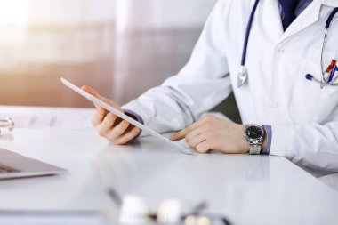 Bilinmeyen erkek doktor, tablet bilgisayarla oturuyor ve çalışıyor. Karanlık bir klinikte, arka planda ışık parıltısı, ellerin yakın çekimi..