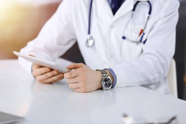 Bilinmeyen erkek doktor, tablet bilgisayarla oturuyor ve çalışıyor. Karanlık bir klinikte, arka planda ışık parıltısı, ellerin yakın çekimi..