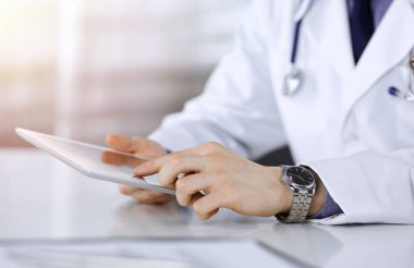 Bilinmeyen erkek doktor, tablet bilgisayarla oturuyor ve çalışıyor. Karanlık bir klinikte, arka planda ışık parıltısı, ellerin yakın çekimi..