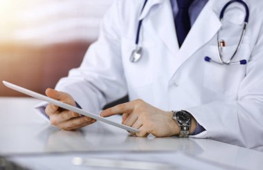 Bilinmeyen erkek doktor, tablet bilgisayarla oturuyor ve çalışıyor. Karanlık bir klinikte, arka planda ışık parıltısı, ellerin yakın çekimi..