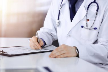 Bilinmeyen bir erkek doktor, karanlık bir klinikte tıbbi geçmişi olan bir panoyla oturuyor ve çalışıyor. Arka planda ışık parıltısı var..