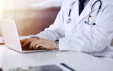 Bilinmeyen bir erkek doktor karanlık bir klinikte dizüstü bilgisayarla oturuyor ve çalışıyor. Arka planda ışık parıltısı var..