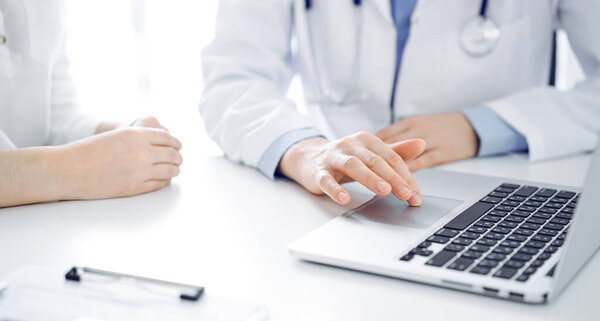 Doctor and patient sitting near each other at the desk in clinic. The focus is on female physicians hands using laptop computer, close up. Medicine concept.