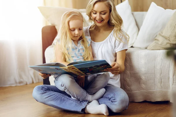 Happy family. Blonde young mother reading a book to her cute daughter while sitting at wooden floor in sunny room. Motherhood concept.