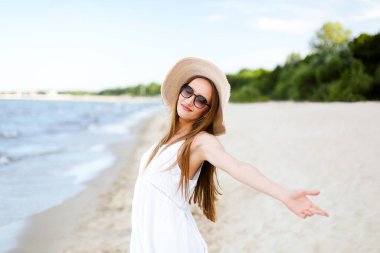 Okyanus sahilinde mutlu, mutlu, mutlu bir kadın şapkası, güneş gözlüğü ve açık elleriyle duruyor. Beyaz yaz elbisesi içinde çok kültürlü bir kadın modelin portresi seyahat sırasında doğanın tadını çıkarıyor.