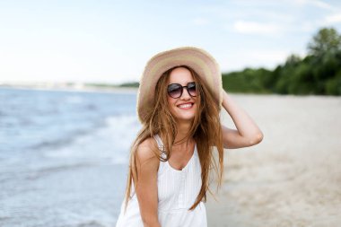 Okyanus sahilinde mutluluk içinde gülümseyen mutlu kadın şapka ve güneş gözlükleriyle poz veriyor. Tatil boyunca doğanın tadını çıkaran beyaz yaz elbiseli bir kadın modelin portresi.