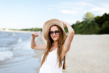 Okyanus sahilinde mutlu, mutlu, mutlu bir kadın şapkası, güneş gözlüğü ve fırfırlı elleriyle duruyor. Beyaz yaz elbisesi içinde doğanın tadını çıkaran çok kültürlü bir kadın modelin portresi.