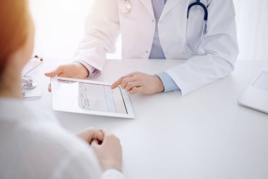 Doktor ve hasta klinikteki masada karşılıklı oturuyorlar. Kadın doktorların elleri tablet bilgisayarın dokunmatik dokunmatik pedini işaret ediyor, kapat. Tıp konsepti.