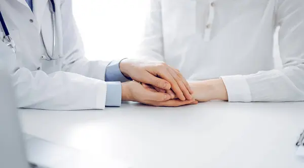 stock image Doctor and patient sitting near each other at the table in clinic office. The focus is on female physicians hands reassuring woman, only hands, close up. Medicine concept.