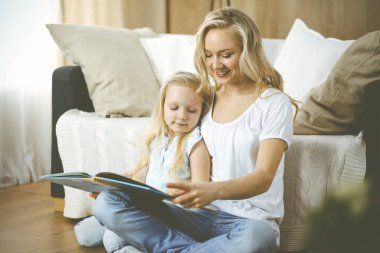 Mutlu bir aile. Sarışın genç anne ahşap zeminde otururken sevimli kızına kitap okuyor. Annelik kavramı.