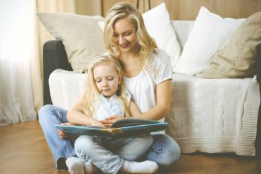Mutlu bir aile. Sarışın genç anne ahşap zeminde otururken sevimli kızına kitap okuyor. Annelik kavramı.