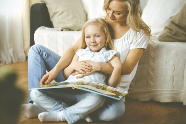 Mutlu bir aile. Sarışın genç anne ahşap zeminde otururken sevimli kızına kitap okuyor. Annelik kavramı.