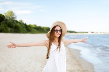 Okyanus sahilinde mutlu, mutlu, mutlu bir kadın şapkası, güneş gözlüğü ve açık elleriyle duruyor. Beyaz yaz elbisesi içinde çok kültürlü bir kadın modelin portresi seyahat sırasında doğanın tadını çıkarıyor.
