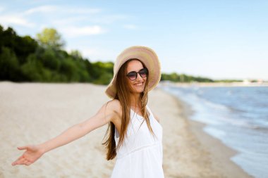 Okyanus sahilinde mutlu, mutlu, mutlu bir kadın şapkası, güneş gözlüğü ve açık elleriyle duruyor. Beyaz yaz elbisesi içinde çok kültürlü bir kadın modelin portresi seyahat sırasında doğanın tadını çıkarıyor.