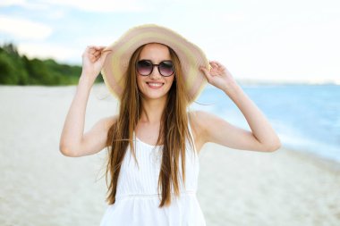 Okyanus sahilinde mutluluk içinde gülümseyen mutlu bir kadının portresi şapkası ve güneş gözlüğü ile duruyor. Beyaz yaz elbisesi içinde seyahat tatilinde doğanın tadını çıkaran bir kadın manken.