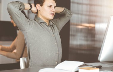 Yeşil tişörtlü, kahverengi kıvırcık saçlı genç işadamı güneşli ofiste bilgisayarla sıkı çalıştıktan sonra ellerini uzatıyor..