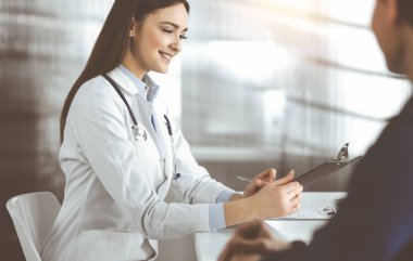 Dost canlısı genç bayan doktor, güneşli klinikte birlikte otururken hastasını dinliyor. Tıp ve sağlık hizmetleri kavramları.