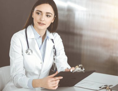 Dost canlısı genç bayan doktor, güneşli klinikte birlikte otururken hastasını dinliyor. Tıp ve sağlık hizmetleri kavramları.